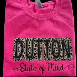 May include: A bright pink sweatshirt with the word "DUTTON" in large, leopard print letters outlined in black. Below the word is the phrase "State of Mind" in a cursive font. The sweatshirt is folded neatly.