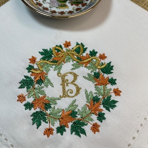 May include: White linen handkerchief featuring an embroidered wreath of green and orange leaves, a gold ribbon, and a gold letter "B". A small porcelain dish with a floral design is in the background.