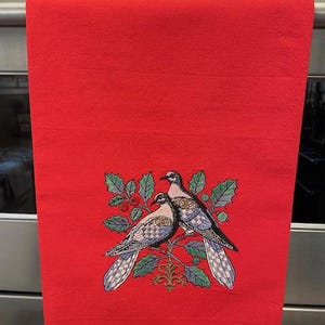 May include: Red kitchen towel with an embroidered design of two white doves with blue and black accents perched on a holly branch with red berries.