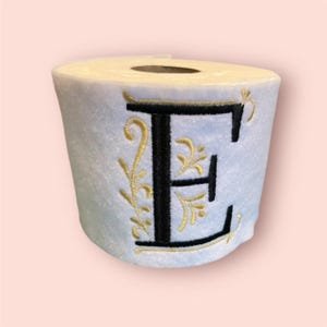 May include: A white toilet paper roll with a black and gold embroidered letter "E" on the front.