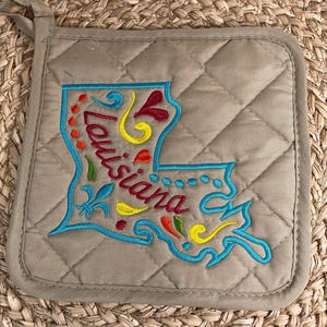 May include: A beige quilted pot holder with an embroidered outline of the state of Louisiana. The embroidery is in turquoise, red, yellow, and green. The word "Louisiana" is embroidered in red and yellow.