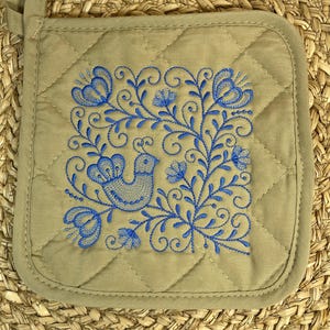 以下が含まれることがあります： 鳥と花の要素が描かれた青い刺繍デザインの、タン色のキルティング鍋つかみ。正方形の鍋つかみには吊り下げ用のループがあり、織り込まれた背景に配置されています。