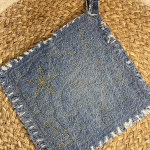 May include: A square denim pot holder with a frayed edge and a loop for hanging. The blue denim is embellished with yellow stitching in a starfish and wave pattern. The pot holder is sitting on a woven straw mat.