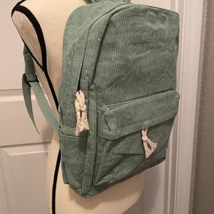 Puede incluir: Una mochila de pana verde con detalles de cuerda blanca. La mochila tiene un bolsillo frontal y correas ajustables.