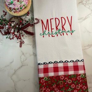 May include: White Christmas kitchen towel with "MERRY CHRISTMAS" embroidered in red and green. The bottom features a red and white gingham pattern and a red candy cane print. A festive holiday decoration.