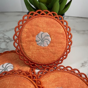 Peut inclure: Ensemble de quatre dessous-de-verre en tissu orange avec un motif en spirale blanc et gris au centre. Chaque dessous-de-verre a un bord festonné décoratif.
