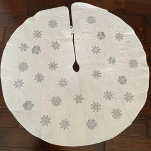 May include: White Christmas tree skirt with a snowflake pattern. The skirt is round with a slit up the middle and a tie closure. The snowflakes are silver and scattered across the surface.