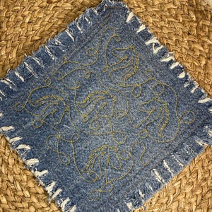 Puede incluir: Salvamanteles cuadrado de mezclilla azul con un lazo para colgar. La mezclilla está adornada con costuras doradas en un patrón arremolinado. Los bordes están deshilachados con hilos blancos.