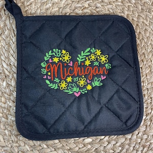 Op de afbeelding: Zwarte, gestikte ovenwant met een hartvormig ontwerp met een krans van bloemen en het woord "Michigan" in oranje borduurwerk.