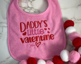 May include: Pink baby bib with the embroidered text "Daddy's little valentine" with a red heart and arrow. The bib is next to a garland of pink, red, and white pom-poms.