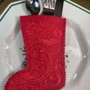 May include: Red felt Christmas stocking with a white embroidered design and the word "NOEL" on the bottom. The stocking is holding a fork, knife, and spoon.