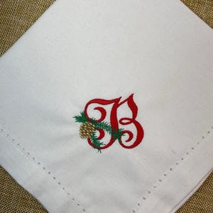 Puede incluir: Servilleta de lino blanco con un monograma bordado en rojo con las letras "SB" con un diseño de piña y ramita de hoja perenne. La servilleta tiene un borde cosido decorativo y está colocada sobre un fondo dorado.