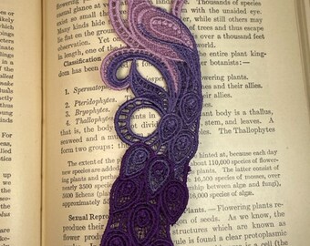 May include: A purple and pink lace bookmark with a floral design. The bookmark is placed on a page of a book with text about flowerless plants.