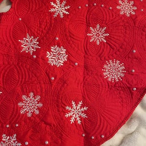May include: A red Christmas tree skirt with white embroidered snowflakes and small pearl accents. The skirt has a quilted pattern and a scalloped edge, creating a festive holiday decoration.
