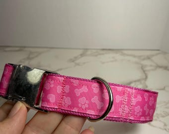 May include: A pink dog collar with a silver buckle and a silver D-ring. The collar is decorated with a white paw print pattern.