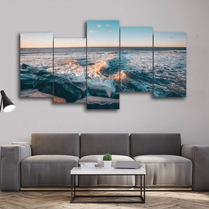 Puede incluir: Una impresión en lienzo de cinco paneles que representa un paisaje marítimo con olas rompiendo y una costa rocosa al atardecer. El océano es de un azul profundo con olas espumosas blancas. Las rocas son de color gris oscuro y húmedas.