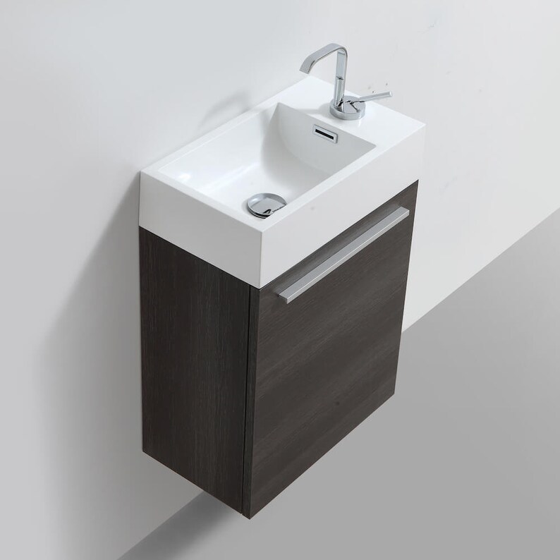 Puede incluir: Un peque&ntilde;o tocador de ba&ntilde;o rectangular blanco con un lavabo simple y un grifo cromado. El tocador tiene un gabinete de madera gris oscuro con una manija plateada.