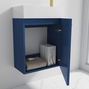 Puede incluir: Un mueble de ba&ntilde;o azul con una encimera blanca y una sola puerta abierta, que revela un estante con una toalla, un tubo de crema y dos cajas de perfume. El mueble tiene una manija dorada.