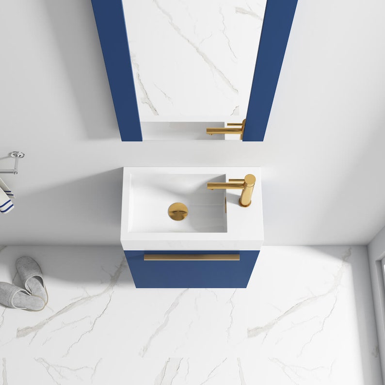 Puede incluir: Un peque&ntilde;o lavabo rectangular blanco con un grifo y un desag&uuml;e dorados. El lavabo est&aacute; montado en un gabinete azul con una manija dorada.