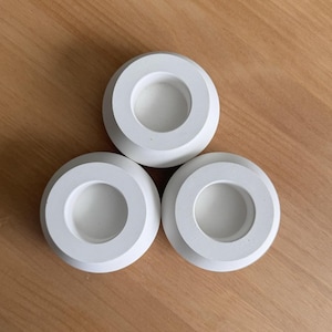 May include: Three white, circular candle holders arranged on a wooden surface. Each holder has a raised outer ring and a recessed center, creating a modern, minimalist design. The holders are made of a solid, opaque material.
