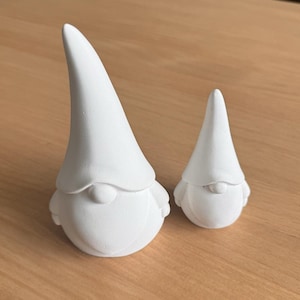 May include: Two white ceramic gnomes, one larger than the other, with pointy hats and rounded bodies. The gnomes are sitting on a wooden surface.
