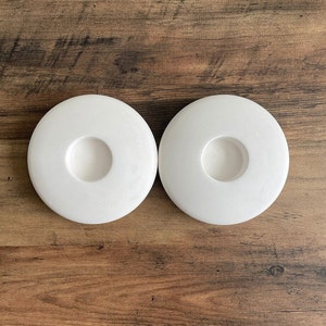 May include: Two white ceramic candle holders with a circular base and a central hole for a candle.