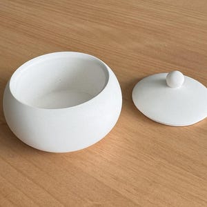 May include: A white ceramic bowl with a lid. The bowl is round and has a smooth, matte finish. The lid has a small knob on top.