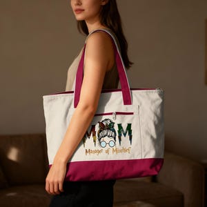 May include: White canvas tote bag with a burgundy bottom and handles. The bag features a front pocket and a zipper closure. The front of the bag has the word "MOM" with a graphic of a woman's face and the words "Manager of Mischief".