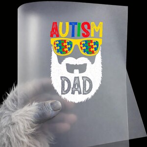May include: A transparent sheet featuring a graphic of a bearded face with sunglasses. The sunglasses are filled with a colorful puzzle pattern. The word "AUTISM" is above the sunglasses in red, yellow, and blue. The word "DAD" is below the beard.