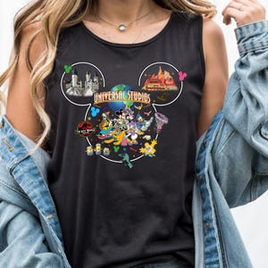 Puede incluir: Camiseta sin mangas negra con un diseño de cabeza de Mickey Mouse y las palabras "Universal Studios". El diseño incluye varios personajes de dibujos animados y atracciones de Universal Studios, como el logotipo de Jurassic Park y los Simpsons.