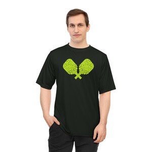 May include: Black t-shirt featuring a lime green graphic of two crossed pickleball paddles. The paddles are made up of small circles. The shirt is short-sleeved and worn by a person.