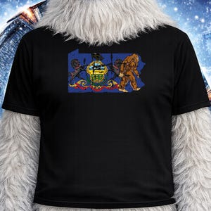May include: Black t-shirt featuring a graphic of the Pennsylvania state seal and a Bigfoot illustration. The design includes the state's coat of arms and a brown Bigfoot walking across the state's outline. The shirt is made of a soft, dark fabric.