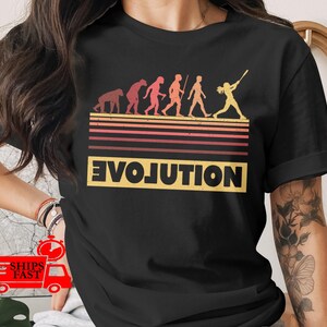 May include: Black t-shirt featuring a retro-style graphic of human evolution, culminating in a baseball player. The word "EVOLUTION" is displayed in a yellow, blocky font. The design includes horizontal stripes in shades of orange and red.