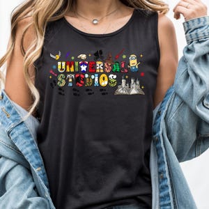 Puede incluir: Camiseta sin mangas negra con las palabras "UNIVERSAL STUDIOS" en letras coloridas de estilo caricatura. El diseño incluye varios iconos temáticos, como un Minion, un sombrero de mago y una bicicleta voladora, con una ilustración de castillo en la parte inferior.