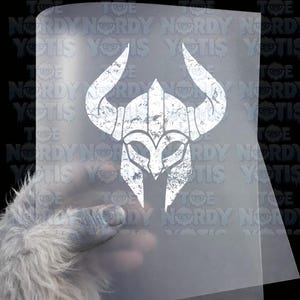 May include: A white, distressed-style graphic of a horned helmet printed on a clear, rectangular transfer sheet. The design features a stylized warrior helmet with two prominent horns, rendered in a weathered, textured white ink finish.