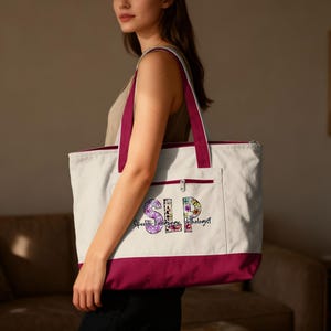 May include: A white tote bag with a burgundy bottom and straps. The bag features a front pocket and a zipper closure. The letters "SLP" are displayed in a floral design, with the words "Speech Language Pathologist" below.