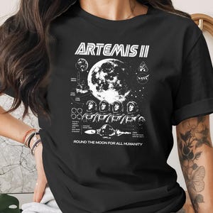 May include: Black t-shirt featuring a white graphic design for the Artemis II mission. The design includes the mission name, a large moon illustration, astronauts, and the text "ROUND THE MOON FOR ALL HUMANITY."