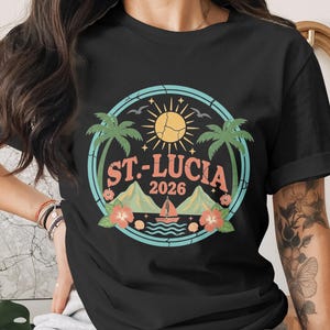 May include: Black t-shirt featuring a circular design with the text "ST. LUCIA 2026". The design includes palm trees, a sun, mountains, a sailboat, and flowers. The shirt is a classic crew neck.