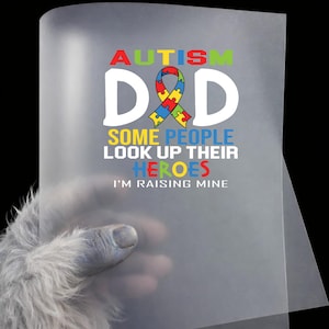 May include: A translucent sheet with the words "AUTISM DAD" in colorful letters, with a puzzle ribbon graphic. Below, the text reads "SOME PEOPLE LOOK UP THEIR HEROES I'M RAISING MINE."