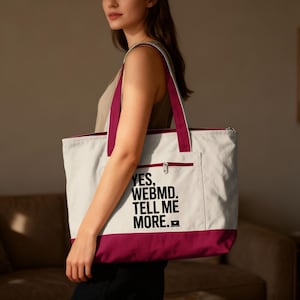 May include: A canvas tote bag with a white body, a deep burgundy bottom, and matching burgundy straps. The bag has a zippered top and a front pocket with the text "YES, WEBMD. TELL ME MORE." printed in black.
