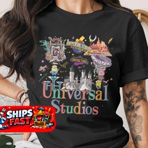 May include: Black t-shirt featuring a colorful graphic of Universal Studios Florida and Hollywood attractions. The design includes cartoon characters, logos, and the words "Universal Studios" in a rainbow font. The shirt is short-sleeved.