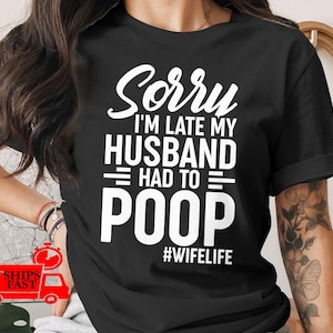 May include: Black t-shirt with the text "Sorry I'm late my husband had to poop #wifelife" in white. The shirt has short sleeves and a crew neck. The text is in a bold, sans-serif font.