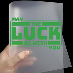 May include: A translucent sheet with the green text "MAY THE LUCK BE WITH YOU" in a rectangular design. The sheet is held by a hand against a black background.