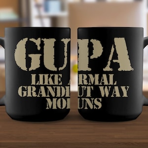 Pode incluir: Caneca de cerâmica preta com o texto "GUPA LIKE A NORMAL GRANDPA BUT WAY COOL". O texto está em uma fonte desgastada, bege. A caneca tem uma alça grande e é exibida em uma superfície de madeira.