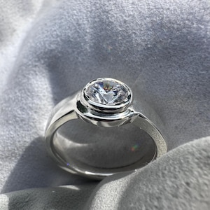 May include: A silver ring with a large, round, clear gemstone. The gemstone is set in a bezel setting, and the band is smooth and polished. The ring is displayed on a soft, gray surface.