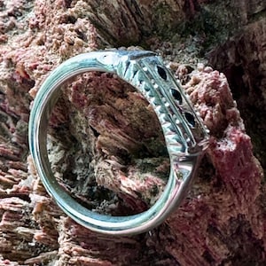 May include: A silver ring with a rectangular top and three small black stones. The ring has a detailed design along the band and is set against a textured, reddish-brown background.