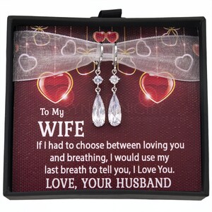 May include: A black gift box with a red and white heart design. The box contains a pair of silver teardrop earrings with clear cubic zirconia stones. The box has a message that reads "To My Wife If I had to choose between loving you and breathing, I would use my last breath to tell you, I Love You. LOVE, YOUR HUSBAND."