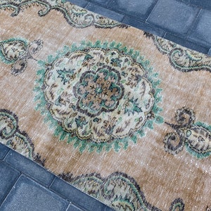 May include: A vintage rug with a beige background and a central medallion in shades of green, brown, and white. The rug has a distressed look and features intricate floral patterns.