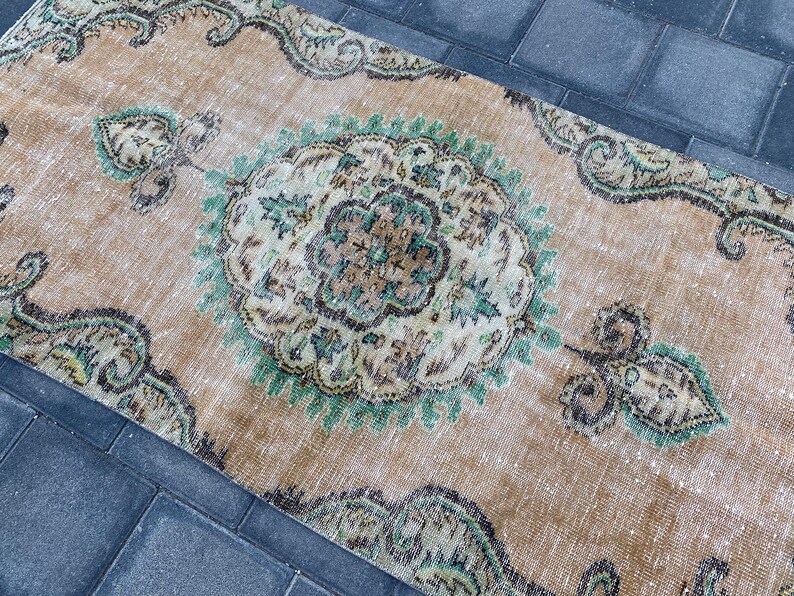 May include: A vintage rug with a beige background and a central medallion in shades of green, brown, and white. The rug has a distressed look and features intricate floral patterns.