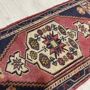 Puede incluir: Una alfombra rectangular que presenta un medall&oacute;n central de color crema con motivos florales geom&eacute;tricos en rojo, amarillo y azul oscuro. La alfombra tiene un borde azul marino y un campo rojo, con complejos patrones tejidos.